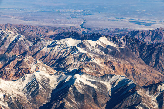 A high-angle aerial shot shows rugged snow-dusted mountain ranges extending to distant flat plains under a clear sky.
