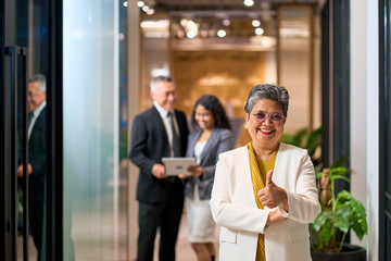 Successful Senior Asian Businesswoman Giving Thumbs Up in Office with Diverse Professional Team, Leadership and Corporate Achievement Concept. © bank215