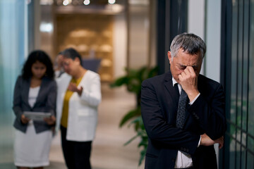 Stressed Senior Asian Businessman with Headache in Office Corridor, Disappointed Male Executive Suffering from Burnout or Business Failure Concept © bank215