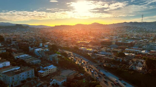 Sunset light over hillside neighborhoods, San Francisco, California. Golden hour aerial view of dense rooftops, a busy freeway and rolling hills silhouetted against the sky.
