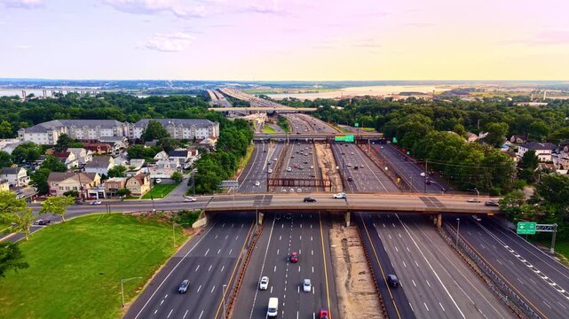 Wide aerial view of freeway overpass at sunset, New Jersey. Residential neighborhood, distant Governor Alfred E Driscoll bridge and traffic under pastel sky. Garden state parkway