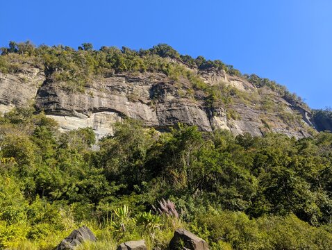 High rocky cliff face covered in lush green forest near Tindu along the Sangu River in Bandarban, under a clear blue sky.