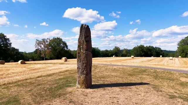 Pr&auml;historischer Gollenstein-Menhir auf einer Wiese bei sommerlicher Erntezeit mit Strohballen unter blauem Wolkenhimmel. Altes Kulturdenkmal in Mitteleuropa.