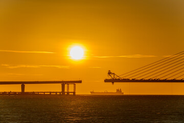 Fototapeta premium A scenic sunset view of the Danjiang Bridge under construction in Tamsui, New Taipei City, Taiwan, with a cargo ship passing by in the distance.