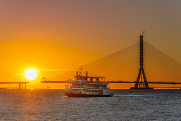 Fototapeta premium A scenic sunset view featuring a passenger ferry boat sailing near the construction site of the Danjiang Bridge in Tamsui, New Taipei City, Taiwan.