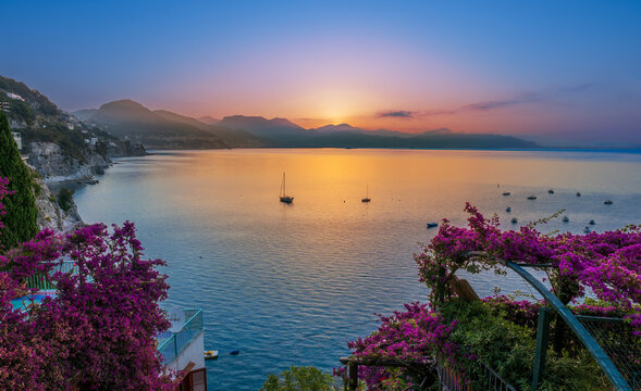Sunrise over Amalfi Coast with magenta bougainvillea flowers framing sea and sailboats in Cetara Italy
