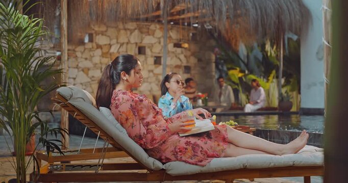 Indian young adult relax woman sitting on sun lounger chair read new novel book enjoy trip day female hold glass drink juice do self care girl child eat fruits spend time have fun joy pool edge place