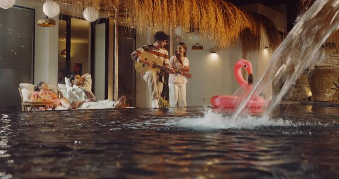 Indian happy young adult relax man male female lying on sun lounger chair enjoy picnic trip night boy hold guitar play music girl sing song clap hand spend time have fun joy at villa pool edge place