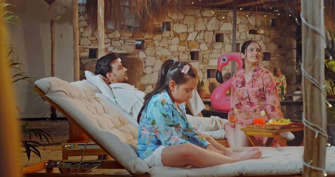 Indian young adult relax man woman girl child sitting on sun lounger talk speak share gossip enjoy trip male female hold glass hold glass drink juice eat fruit spend time have fun joy villa pool edge