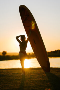 Woman standing by a lake at sunset while holding a large surfboard upright on the grassy shore area
