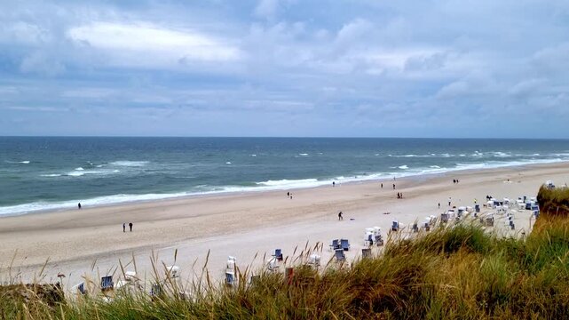 Scenic coastal view of sandy beach with people walking along the shore, beach chairs arranged on the sand, and gentle waves lapping at the shoreline