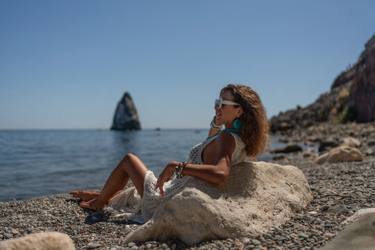 Caucasian woman wearing white lace dress poses on pebble beach against vast sea under bright daylight