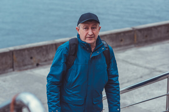 Thoughtful senior man aged 50s 60s in blue jacket and cap standing alone outdoors looking into distance, psychological mental health concept of loneliness and reflection