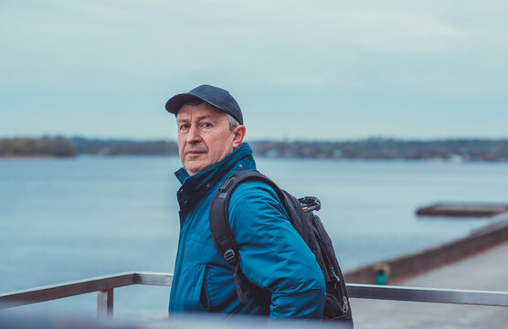 Thoughtful senior man aged 50s 60s in blue jacket and cap standing alone outdoors looking into distance, psychological mental health concept of loneliness and reflection
