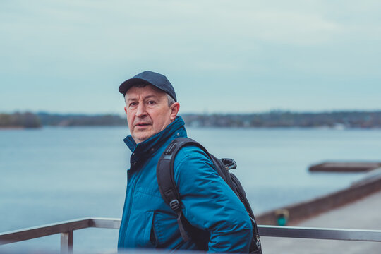 Thoughtful senior man aged 50s 60s in blue jacket and cap standing alone outdoors looking into distance, psychological mental health concept of loneliness and reflection
