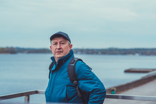 Thoughtful senior man aged 50s 60s in blue jacket and cap standing alone outdoors looking into distance, psychological mental health concept of loneliness and reflection