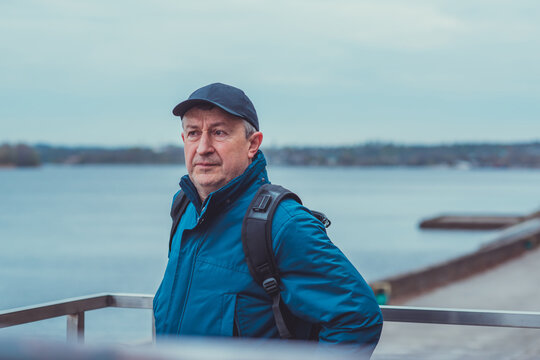 Thoughtful senior man aged 50s 60s in blue jacket and cap standing alone outdoors looking into distance, psychological mental health concept of loneliness and reflection
