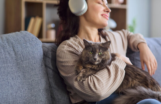 Woman with cat in headphones listening music. Closeup at home on a sofa shows her cuddling pet, smiling and enjoying wireless audio, creating a relaxing cozy mood. Cozy calm home pet mood.