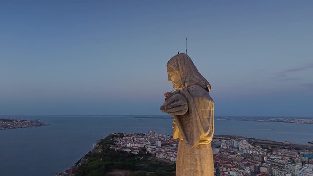 Close aerial side view of the illuminated Cristo Rei monument overlooking Almada and the Tagus estuary in Portugal beneath a deep blue evening sky.