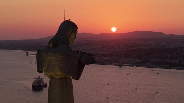 Aerial view of the Cristo Rei monument overlooking the Tagus River, sailboats, and distant hills beneath a vivid orange sunset near Almada, Portugal.