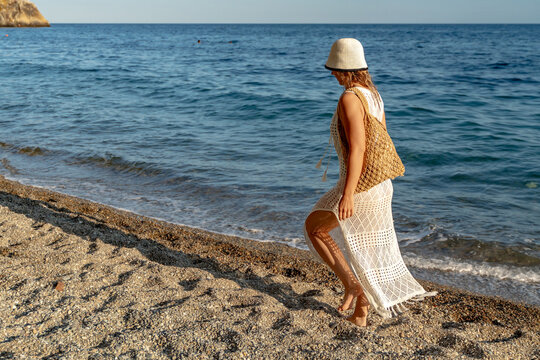Summer leisure young woman wearing crochet dress and sun hat walking along pebble beach near blue ocean waves