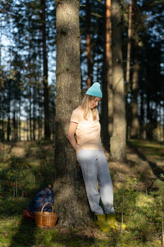 Tranquil smiling woman forager backpacker taking moment to enjoy harmony of nature in forest, calm relaxed female leaning against tree in summer woodlands during mushroom picking hobby activity.