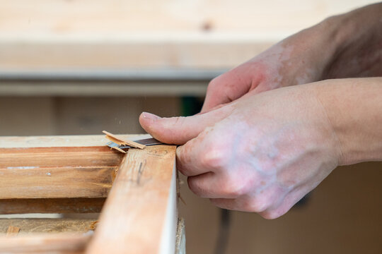 Woodworker holding chisel tool and carving layers of old wood from vintage window frame in workshop, male master using professional carpentry instrument for repairing and restoring antique furniture.
