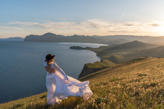 Woman hat dress, woman standing on a grassy hill overlooking the ocean at sunset enjoying nature and freedom during travel