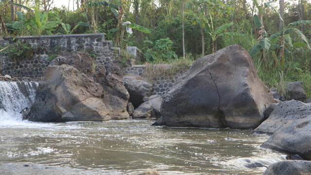 A scenic outdoor landscape photograph capturing large natural boulders situated in front of a small river weir waterfall dam.