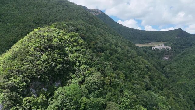 Montagna ricoperta da foresta verde con pendii ripidi, zona rocciosa vicino alla cima e cielo azzurro con nuvole bianche. Paesaggio naturale ampio, luminoso e incontaminato
