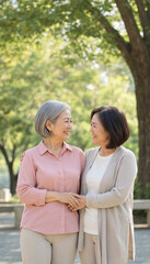 Fototapeta premium Two middle aged women smiling and holding hands in a park, showing friendship and warmth. They stand close together under trees with soft natural light.