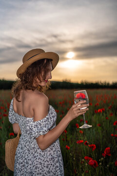 Summer evening portrait of young woman wearing straw hat holding glass with Papaver rhoeas flower in blooming poppy field