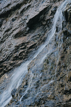 A tight vertical crop of a thin waterfall streak trickling down a dark, textured rock face.