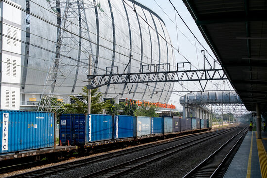 Jakarta, Indonesia - July 29, 2026: Container freight train passes JIS Station beneath a pedestrian skybridge with Jakarta International Stadium in the background.