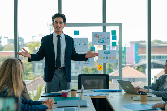 Professional speaker presents business strategy beside infographic board. Colleagues review metrics, compare charts, discuss planning, exchange insight, improve teamwork, performance.