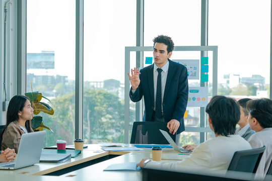 Professional speaker explains business strategy using presentation board while colleagues discuss objectives, compare analytics, review metrics, improve communication, teamwork, leadership.