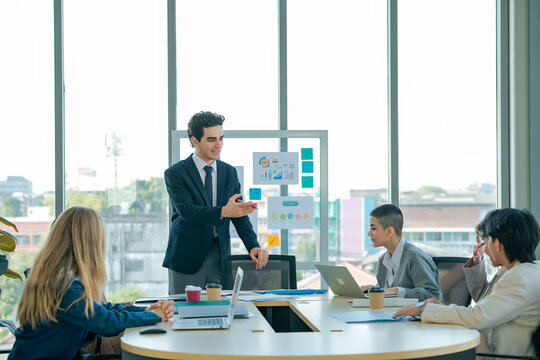 Business manager explains strategy during meeting while colleagues review presentation, exchange ideas, analyze performance, discuss solutions, improve communication, planning, teamwork.