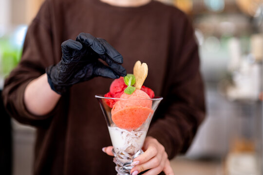 Gloved hands place mint sprig on strawberry and raspberry ice cream parfait at cafe.