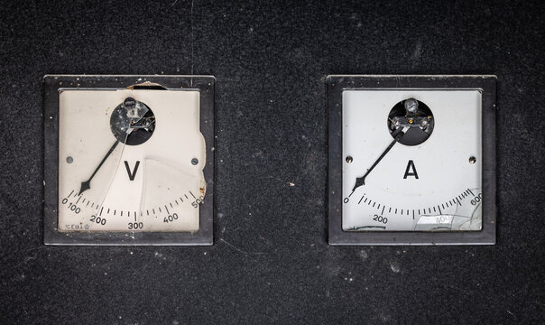 Old analog voltmeter and ammeter installed on an industrial electrical control panel with weathered surfaces and damaged glass.