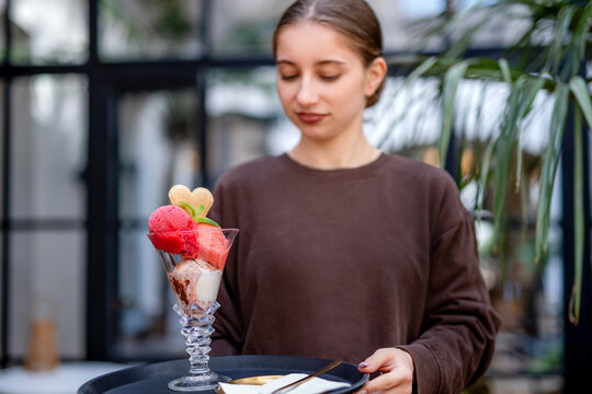 Waitress carries tall glass sundae with red and white scoops, cookie, spoon on tray.