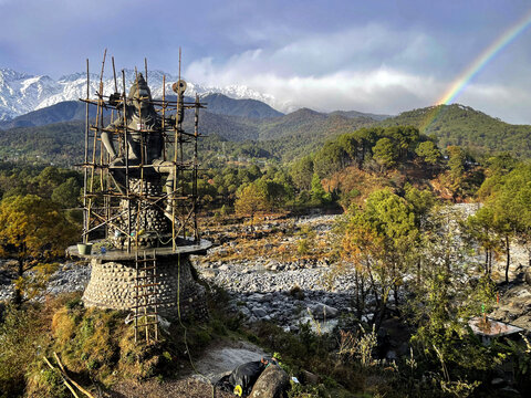 A statue of Shiva under construction in Palampur, India, with mountains and a rainbow.