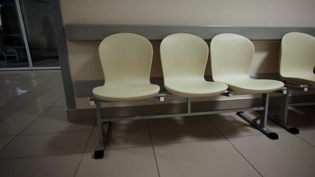 Empty waiting chairs stand lined up along corridor wall near glass doors. Dim indoor lighting creates slightly somber evening atmosphere