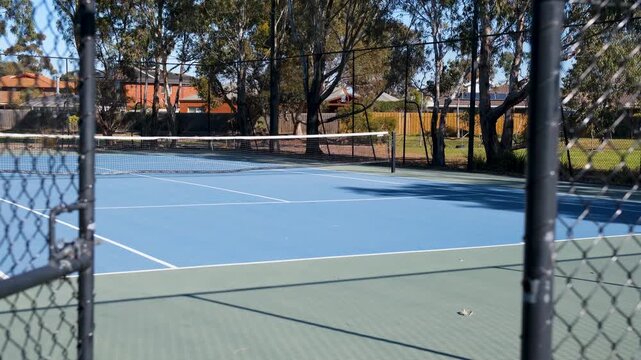 A blue hardcourt suburban tennis court framed through an open chain-link fence in a local park area in Melbourne, Australia,&nbsp;bordered by mature eucalyptus trees and residential houses