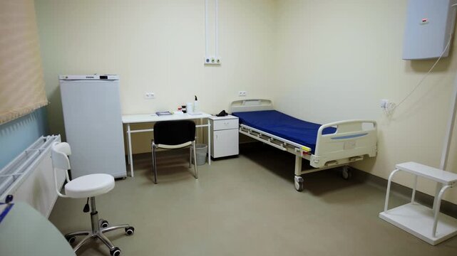 Medical inpatient room features adjustable hospital bed small refrigerator and office chair. Ordered setup reflects healthcare facility