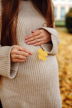 Pregnant woman wearing beige sweater holding yellow leaf autumn season outdoors. Fall maternity photoshoot, pregnancy concept, cozy motherhood waiting, nature park lifestyle, October pregnancy