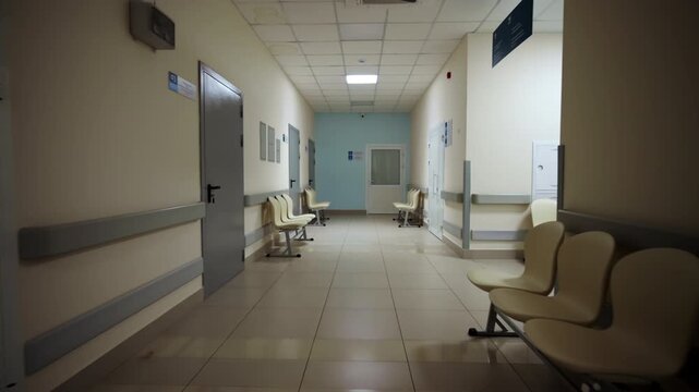 Wide hospital corridor shows beige plastic chairs beside closed doors. Bright ceiling lighting fills modern institutional hallway setup
