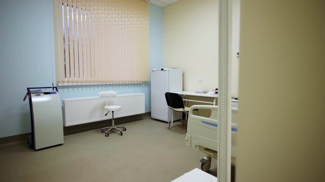 Medical office contains rolling clinical device and vertical window blinds. Clean setup indicates prepared healthcare space for patients
