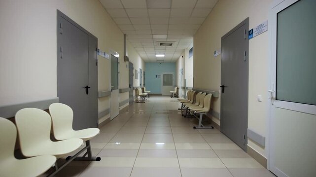 Long hospital corridor features rows of empty waiting chairs lining beige walls. Overhead square lamps illuminate spacious clinical hallway