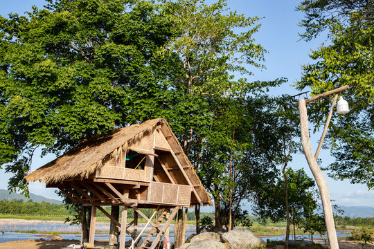 A traditional rustic bamboo hut with a thatched roof elevated on stilts, surrounded by lush green trees near a scenic lake under a clear blue sky.