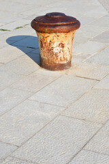 Naklejka premium A single old rusty iron mooring bollard stands firmly on a paved stone pier under bright natural sunlight